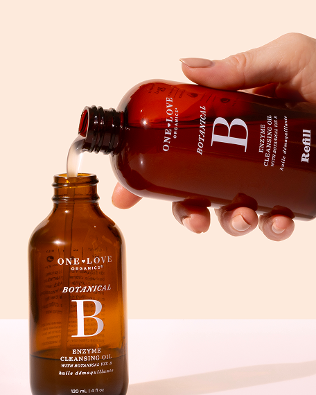 A hand pours clear liquid from a brown amber bottle labeled One Love Organics Botanical B Refill into a matching empty bottle against a warm cream background.