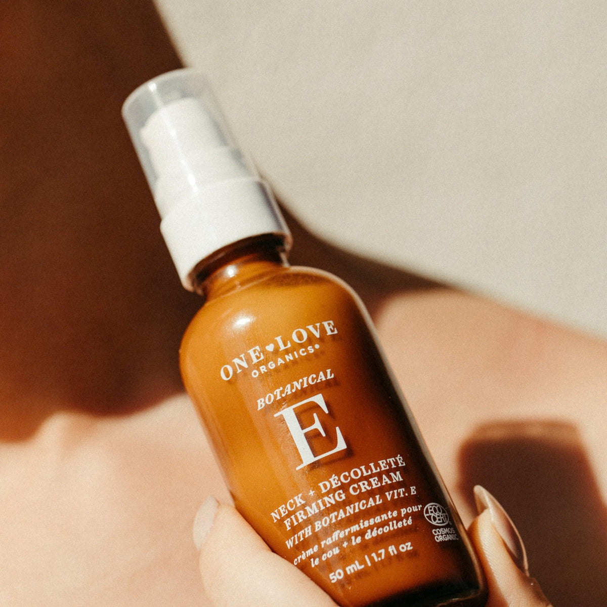 A hand holding a brown amber spray bottle of One Love Organics Botanical E Neck & décolleté Firming Cream against a warm, sunlit beige background.
