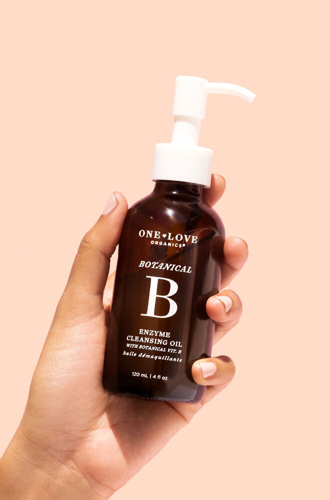 A hand holding a dark amber bottle of One+Love Organics Botanical B Enzyme Cleansing Oil